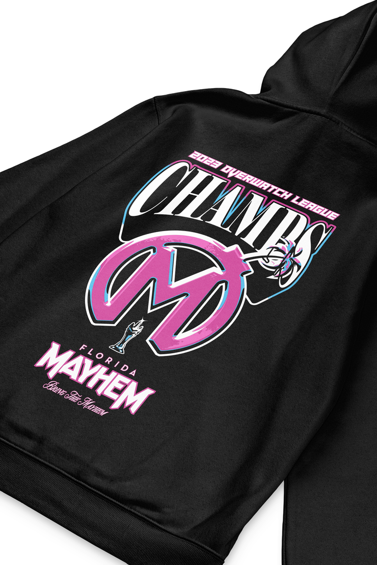 A close-up of a black hoodie showcasing vibrant print details. On it, the bold pink and blue text "2023 Overwatch League CHAMPS" stands out prominently. Beneath the text is a large stylized logo with the initials "FM" in a gradient of pink and blue, representing the "Florida Mayhem" team. Below the logo is the word "MAYHEM" in pink, and a smaller depiction of a character with a trophy, accompanied by the words "Florida Mayhem - Born For Mayhem" in pink and white.