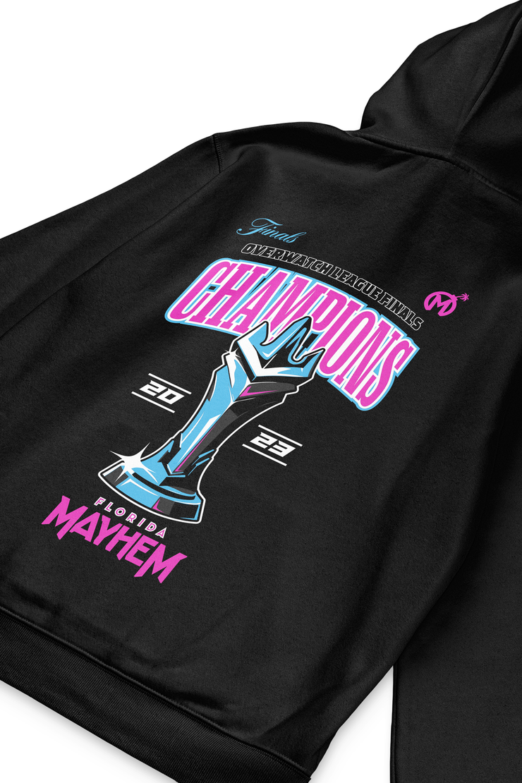A clos up of A black pullover hoodie with a design celebrating the "Finals Overwatch League Finals." It features the word "CHAMPIONS," a blue and teal trophy illustration, and the years 2023 At the bottom, "Florida Mayhem" is printed in pink with the team&